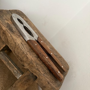 May include: A vintage nutcracker with a stainless steel cracking mechanism and wooden handles. The handles are a dark brown colour and are attached to the metal with small white rivets. The nutcracker is resting on a weathered wooden surface.