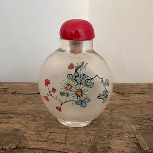 Puede incluir: Pequeña botella de vidrio redonda con tapón rojo. La botella está decorada con un diseño floral en tonos azules, verdes y rojos. La botella está sobre una superficie de madera.