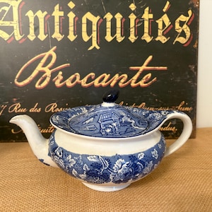 Puede incluir: Una tetera vintage con un diseño floral azul y blanco. La tapa presenta una ilustración detallada. El fondo incluye un letrero que dice "Antiquités Brocante".