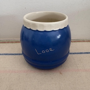 Puede incluir: Un tarro de cerámica azul con un borde blanco y la palabra "Looe" escrita en la parte delantera.