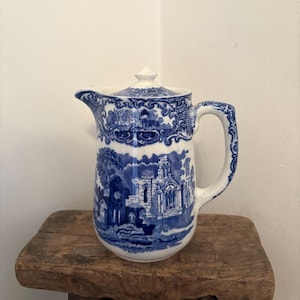 Puede incluir: Jarra de cerámica blanca y azul con tapa, con un diseño detallado de paisaje. La jarra tiene un asa y un pico, y está colocada sobre un pequeño taburete de madera. El diseño está en una combinación de colores azul y blanco clásica.