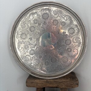 May include: A large, round, silver-coloured metal serving tray featuring an intricate embossed floral and scrollwork pattern across its surface. The tray has a raised, perforated lattice-style border along the outer edge and a smooth centre.