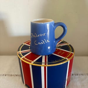 Puede incluir: Una pequeña taza de cerámica azul con asa blanca. La taza tiene las palabras "Belvoir Castle" escritas en dorado en la parte delantera. La taza está sentada sobre una caja con un diseño de Union Jack.