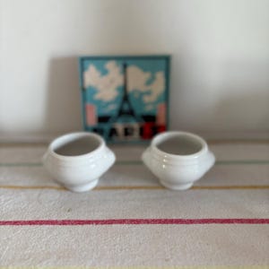 Op de afbeelding: Twee kleine, witte keramische kommen met handvatten worden gepresenteerd op een gestreept tafelkleed. Op de achtergrond is een ingelijste afbeelding van de Eiffeltoren met het woord "PARIS" te zien.