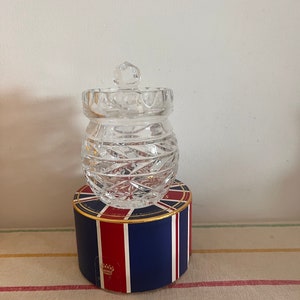 Op de afbeelding: Een glazen pot met deksel, met een geslepen glasontwerp. De pot staat op een blauwe en rode doos met een gouden rand en een kroonlogo.