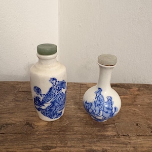 Puede incluir: Dos botellas antiguas de porcelana blanca con escenas pintadas en azul. Cada botella tiene un tapón verde. Una botella es más alta y cilíndrica, mientras que la otra es más corta y redonda. Ambas botellas están sobre una superficie de madera.