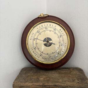 May include: A round analog barometer with a dark wood frame and a gold-toned metal face. The dial features black text including "PLUIE," "VARIABLE," "BEAU," and "TRES SEC," along with "BAROMETRE PRECISION FAIVRE-DUTEC MORTEAU" at the bottom.