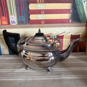 Può includere: Una teiera d'argento con manico nero e tre piedini. La teiera è appoggiata su una tovaglia a righe davanti a una libreria.