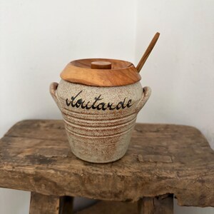 May include: A ceramic mustard pot with a wooden lid and spoon. The pot is beige with the word "Moutarde" written in black script. The lid and spoon are made of light-colored wood. The pot has two small handles.