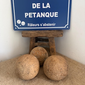 Op de afbeelding: Een rechthoekig blauw metalen bord met witte tekst PLACE DE LA PETANQUE en Râleurs s'abstenir, voorzien van een afbeelding van twee witte petanqueballen. Daaronder liggen drie verweerde, bolvormige houten petanqueballen.