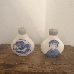Puede incluir: Dos botellas de cerámica blanca con tapones grises. Una botella presenta un diseño de dragón azul, y la otra representa a una persona con atuendo azul. Las botellas se exhiben sobre una superficie de madera rústica.