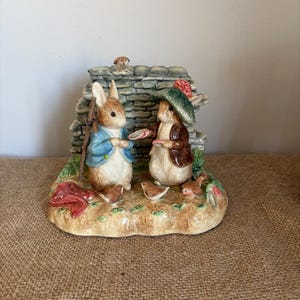 Puede incluir: Figura de cerámica con dos conejos en un jardín. Un conejo lleva una chaqueta azul y un bastón, el otro una chaqueta marrón y un sombrero verde. También hay un muro de piedra y pequeños pájaros.