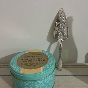 May include: A teal blue tin with a gold trim and a white label that reads "Fortnum & Mason Traditional Dundee Cake". A silver cake server with an ornate design is next to the tin.