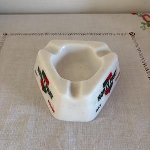 Può includere: Posacenere in ceramica bianca di forma triangolare con un pozzetto centrale. Il posacenere presenta le parole "NOILLY PRAT" in rosso e verde, con "ROUGE" e "DRY" stampati sui lati. Un oggetto vintage.