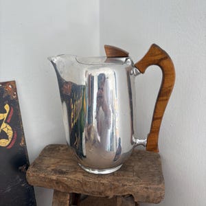 Puede incluir: Una jarra de metal plateado pulido con un asa de madera curvada y un detalle de madera a juego en la tapa. La jarra tiene una superficie lisa y reflectante con un pico prominente y una base resistente, combinando metal y madera.