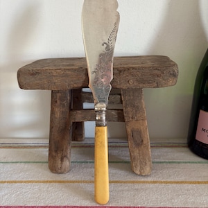 May include: A vintage butter knife with a silver blade and a cream-colored handle. The blade has an ornate floral design. The knife is displayed on a small wooden stool, suggesting a rustic or antique theme.