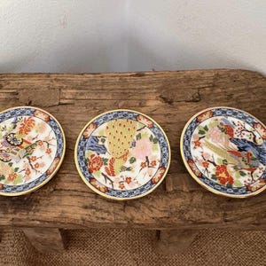 Puede incluir: Tres platos decorativos de cerámica con intrincados diseños florales y de aves. Cada plato presenta un borde dorado, un borde azul y blanco y una imagen central de un pavo real, flores y otros elementos decorativos. Los platos se exhiben sobre una superficie de madera rústica.