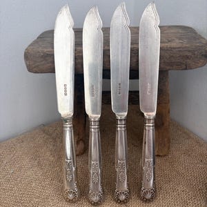 Peut inclure: Un ensemble de quatre couteaux argentés dotés de lames courbes et de manches ornés de motifs en forme de coquille. Chaque lame de couteau porte une série de petits poinçons rectangulaires. Le métal présente une finition polie et détaillée.