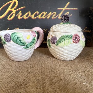 Peut inclure: Un pot à lait et un sucrier avec couvercle en céramique blanche à motif de vannerie. Les deux pièces sont ornées de feuilles vertes, de fleurs roses et de mûres violet foncé en relief. Le pot à lait possède une anse rose pâle.
