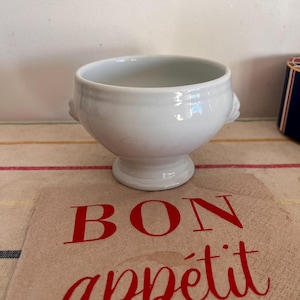 Könnte beinhalten: Weiße Suppenschüssel aus Keramik mit zwei Löwenkopf-Griffen. Die Schüssel steht auf einer braunen Papierserviette mit dem Schriftzug "Bon appétit" in Rot.