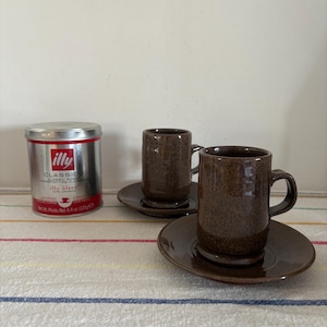 Puede incluir: Dos tazas de espresso de cerámica marrón con platillos a juego sobre un mantel a rayas. Se ve una lata de café Illy plateada. Las tazas tienen un acabado texturizado y asas pequeñas. Los platillos son redondos con un ligero borde.