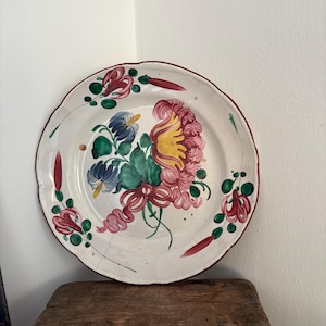Op de afbeelding: Een witte keramische plaat met een geschulpte rand, versierd met een bloemenontwerp in roze, geel, blauw en groen. De rand is rood omlijnd. De plaat staat op een rustieke houten kruk.