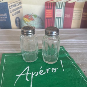Peut inclure: Deux salières et poivrières en verre transparent avec des couvercles argentés sur une serviette verte avec le mot "Apéro!" écrit en blanc.