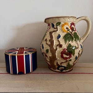 May include: A white ceramic jug with a floral design in shades of red, blue, green, and yellow. The jug has a handle and a round body. The jug is sitting on a striped surface.