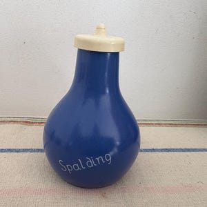 Puede incluir: Una botella de cerámica azul con una tapa blanca y la palabra "Spalding" escrita en la parte delantera. La botella está sobre una superficie rayada blanca y azul.