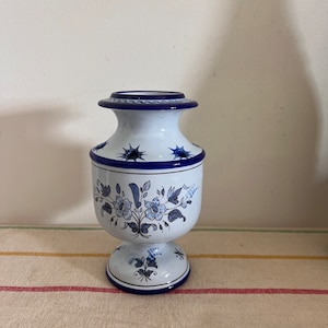 May include: A white ceramic vase with a blue and white floral design. The vase has a round base and a narrow neck. It is decorated with blue stars and a floral pattern.