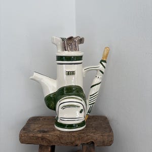 Puede incluir: Una tetera de cerámica con forma de bolsa de golf, con una combinación de colores blanco y verde. La tetera tiene un pico, un asa y un diseño de paraguas de golf. Los palos de golf son visibles en la parte superior. La tetera está sobre un taburete de madera.