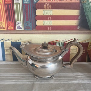Könnte beinhalten: Eine silberne Teekanne mit einem Holzgriff und einem Deckel. Die Teekanne steht auf einer weißen und braunen gestreiften Tischdecke. Die Teekanne hat einen runden Körper und einen Ausguss an der Seite. Die Teekanne steht vor einem Bücherregal mit Büchern in verschiedenen Sprachen.