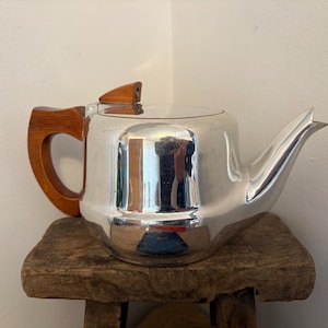 Puede incluir: Una tetera plateada pulida con asa y pomo de tapa de madera. La tetera está sobre un pequeño taburete de madera rústica. La superficie de la tetera refleja el entorno.