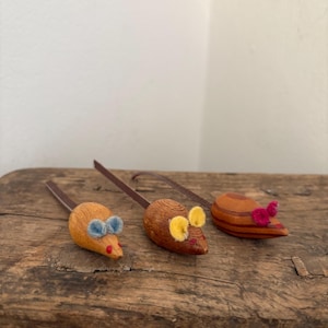 Könnte beinhalten: Drei handgefertigte Spielzeugmäuse mit braunen Lederschwänzen liegen auf einer rustikalen Holzoberfläche. Die Mäuse sind aus Filz gefertigt und haben verschiedenfarbige Akzente: blaue Schleife, gelbe Ohren und rosa Akzente. Die Spielzeuge sind für Katzen.