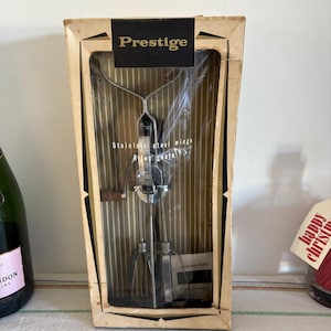 May include: A vintage Prestige egg beater in its original box. The box is beige with black trim and a clear plastic window. The egg beater has stainless steel wings and a wooden handle. The box reads "Prestige" in gold lettering.