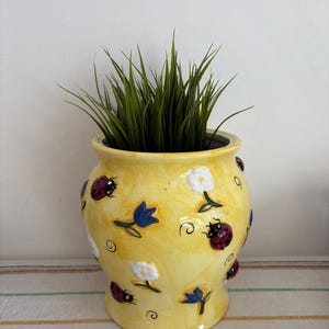 May include: A yellow ceramic planter with a bulbous shape, adorned with ladybugs, blue flowers, and white flowers. The planter contains green grass. The design is whimsical and decorative.