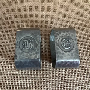 Puede incluir: Dos anillos de servilleta de plata con acabado martillado. Cada anillo tiene un diseño circular con una letra, "G" y "F", en fuente gótica.
