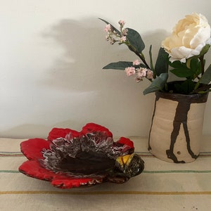 Puede incluir: Un plato de cerámica rojo y marrón con forma de flor con un centro amarillo. El plato está sobre un mantel a rayas.