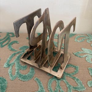Puede incluir: Un portacartas vintage de metal plateado. El soporte tiene una base rectangular con un diseño de rejilla y cuatro letras verticales. Las letras están estilizadas con elementos curvos y angulares. La pieza es probablemente de mediados de siglo.
