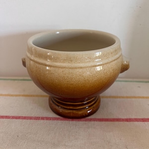 Puede incluir: Un tazón de sopa de cerámica marrón y blanco con dos asas. El tazón tiene una base de pedestal.