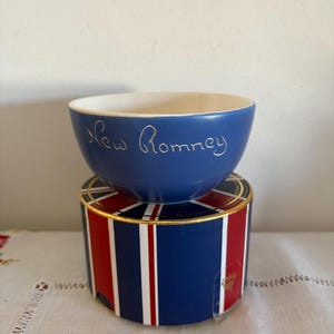 Puede incluir: Un cuenco de cerámica azul con interior blanco y letras doradas que dicen "New Romney". El cuenco está sobre una caja con un diseño a rayas rojas, blancas y azules.