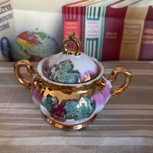 May include: A vintage sugar bowl with a lid, featuring a white base with pink and gold accents. The bowl is adorned with green and purple leaf and berry designs. It has gold handles and a gold finial on the lid.