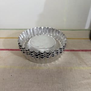May include: A stack of small, silver, fluted tart pans. The pans have a circular base and a wavy, crimped edge. They are made of a shiny, metallic material and are stacked on top of each other.