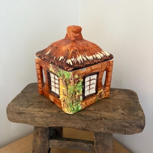 May include: A ceramic cottage-shaped container with a thatched roof and brick walls. The container has two windows with white frames and is decorated with green vines. It sits on a rustic wooden stool.