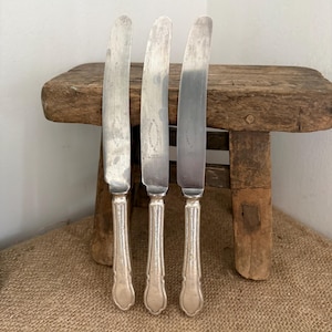 May include: Three silver-toned metal knives with curved blades and ornate, textured handles. Each knife features a distinct oval-shaped hallmark stamped on the blade surface, showcasing a classic design with a polished finish and detailed metalwork.