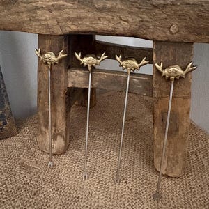May include: Four cocktail forks with crab-shaped gold-coloured tops and long, slender silver stems. The forks are on a burlap surface in front of a rustic wooden bench.