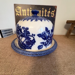 May include: A blue and white ceramic cheese dome with a floral design. The dome sits on a matching plate with a decorative edge. The word "Antiquités" is visible in the background.