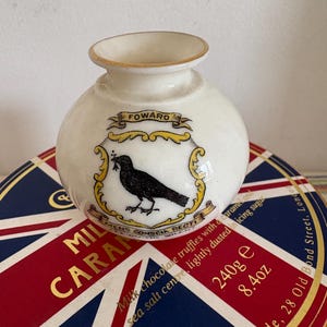 Puede incluir: Un jarrón de cerámica blanca con un diseño de cuervo negro y detalles dorados. El jarrón tiene las palabras "Foward" y "Mens Conscia Recti" escritas en él. El jarrón está sentado encima de una caja con las palabras "Milk Chocolate Truffles with Carame Sea Salt Centre, Lightly Dusted Icing Sugar" y "240g e 8.4oz" escritas en ella.