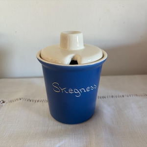 Op de afbeelding: Een blauwe keramische pot met een wit deksel en het woord "Skegness" in wit op de voorkant. De pot heeft een klein gaatje in het deksel om te gieten.