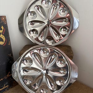 Könnte beinhalten: Zwei runde Servierplatten aus Edelstahl mit integrierten Seitengriffen. Jede Platte verfügt über ein zentrales, blumenartiges Design mit sechs länglichen, blütenblattförmigen Vertiefungen und sechs kleineren kreisförmigen Mulden.